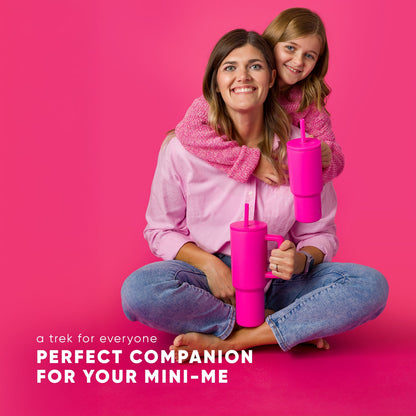 Adult woman and young child seated together on hot pink background, both wearing matching magenta knit sweaters and blue jeans, holding bright fuchsia Simple Modern Trek tumblers with straws. Text reads "a trek for everyone"