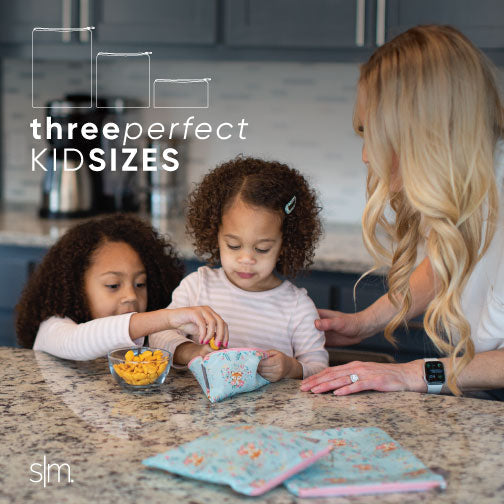 Adult and two children in a modern kitchen with gray cabinetry opening an Ellie Snack Bag, demonstrating the product in a family home setting with neutral-toned décor and clothing.