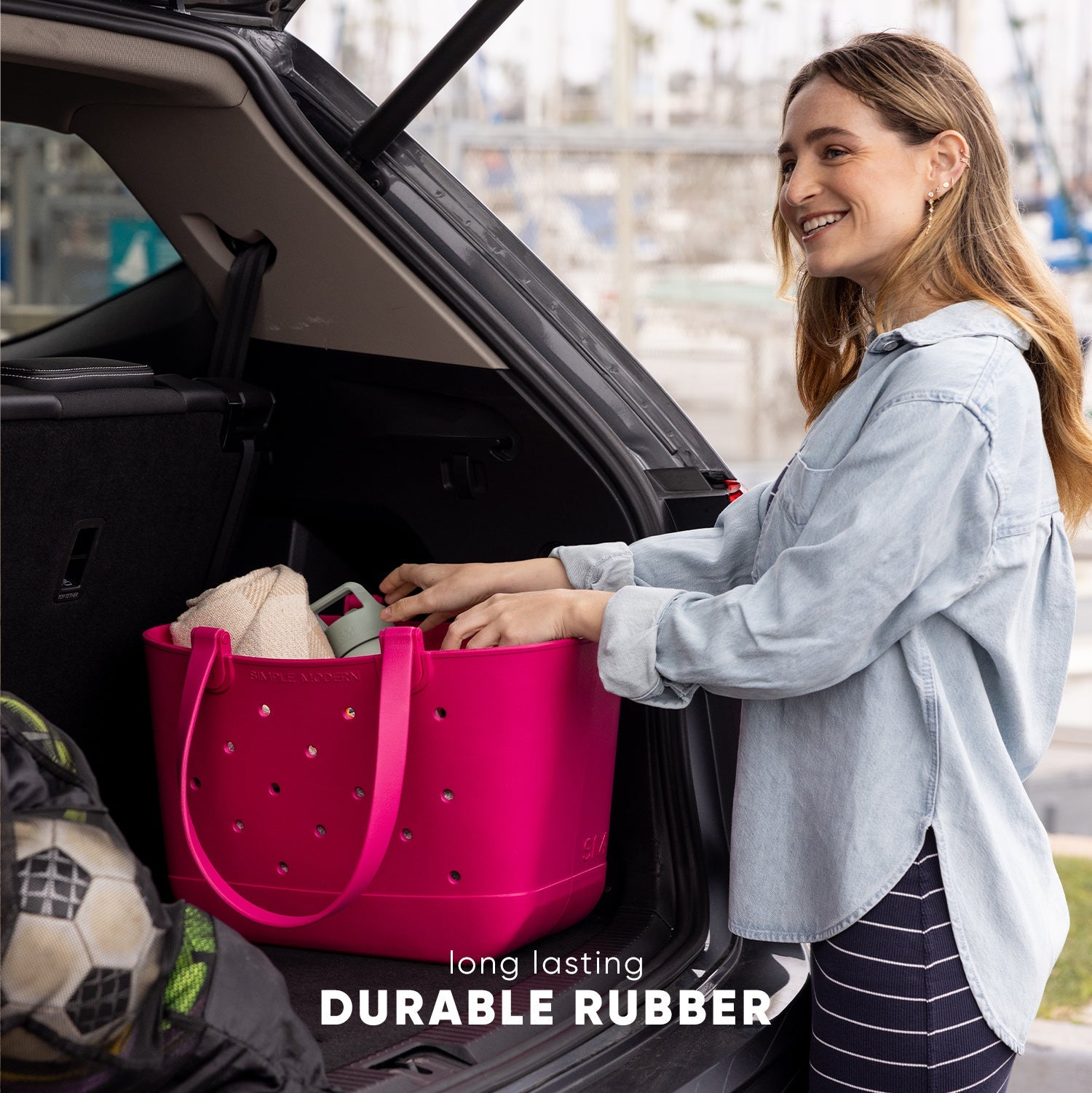 Woman smiling while loading a hot pink rubber tote bag with rope handles into a car trunk. Bag contains folded textiles and packed items. Text reads "long lasting DURABLE RUBBER." Urban cityscape visible in background.
