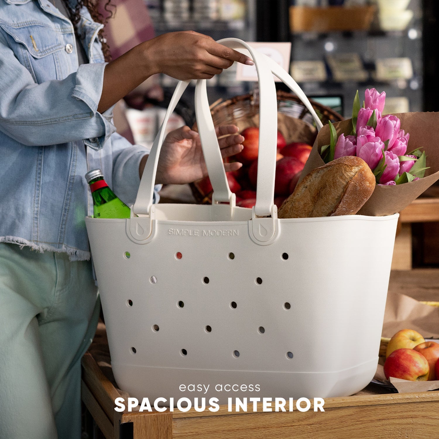White Simple Modern tote bag with perforated ventilation holes filled with fresh groceries including bread, tulips, apples, and tomatoes. Person in blue denim jacket holding bag at wooden market stand. Text overlay reads