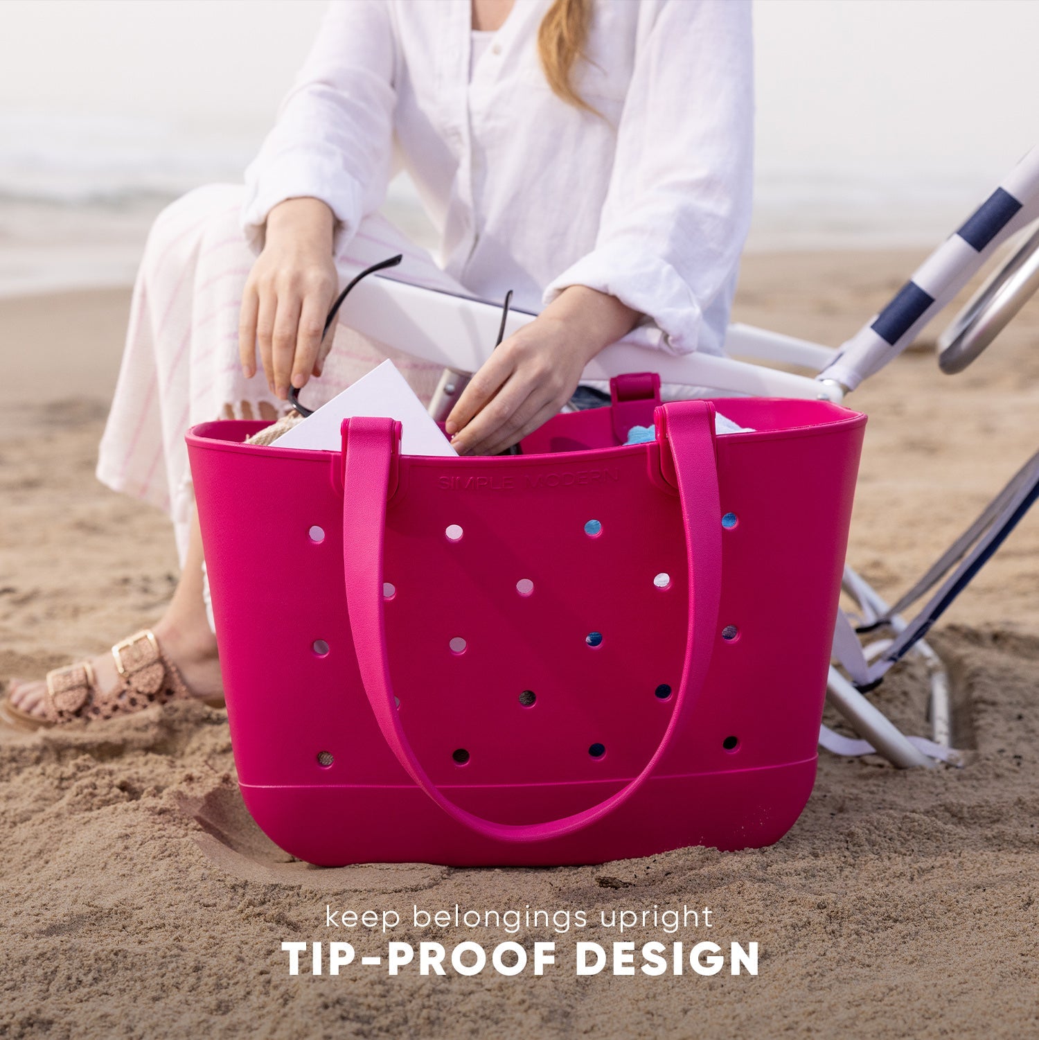 Bright magenta Simple Modern Getaway Bag with perforated design held by woman in white linen on beach. Text reads "keep belongings upright TIP-PROOF DESIGN." Beach chairs and ocean visible in background.