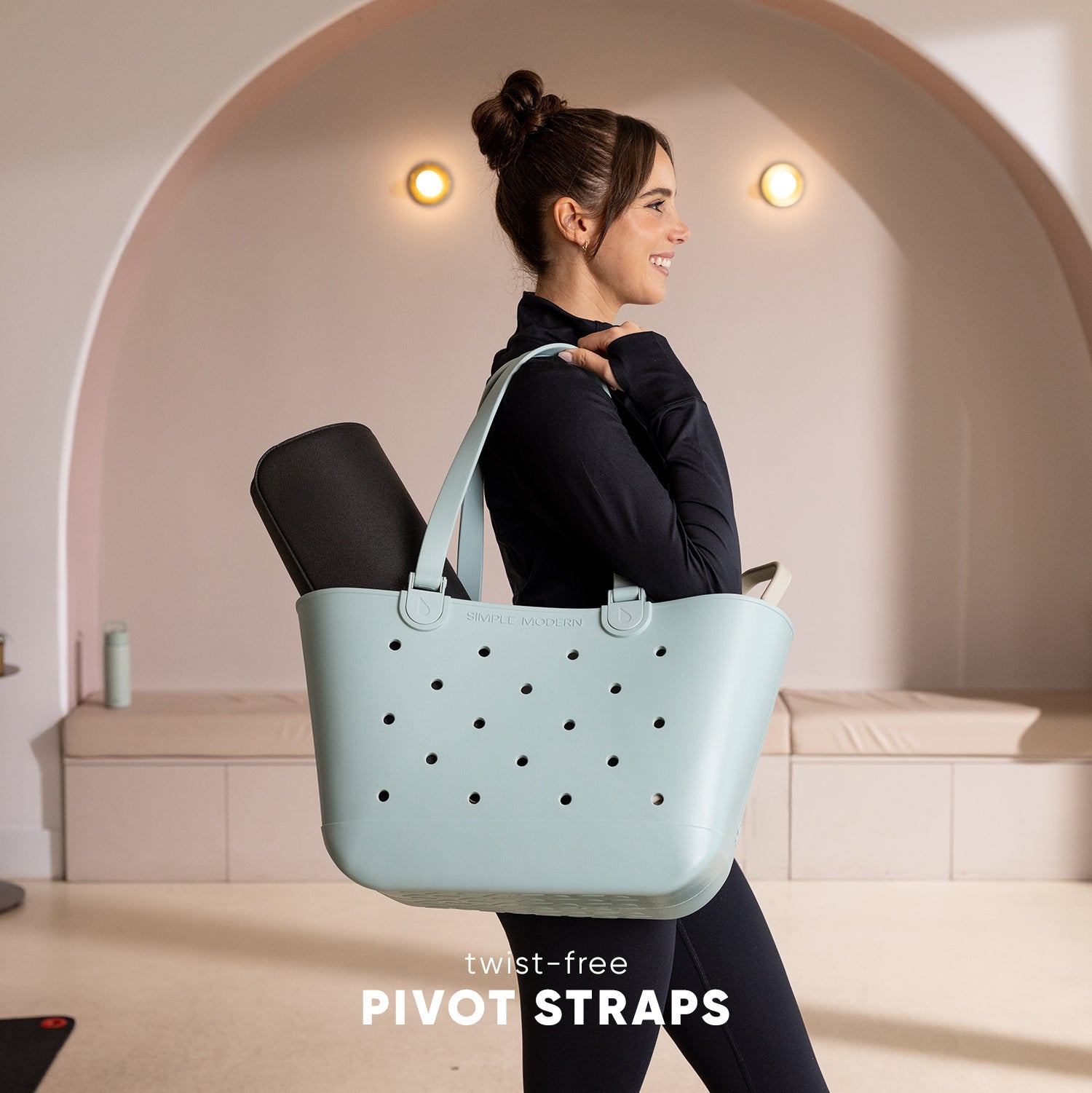 Woman in black athletic wear walking while carrying a pale blue-gray perforated tote bag with twist-free pivot straps, in a minimalist studio setting with warm lighting.