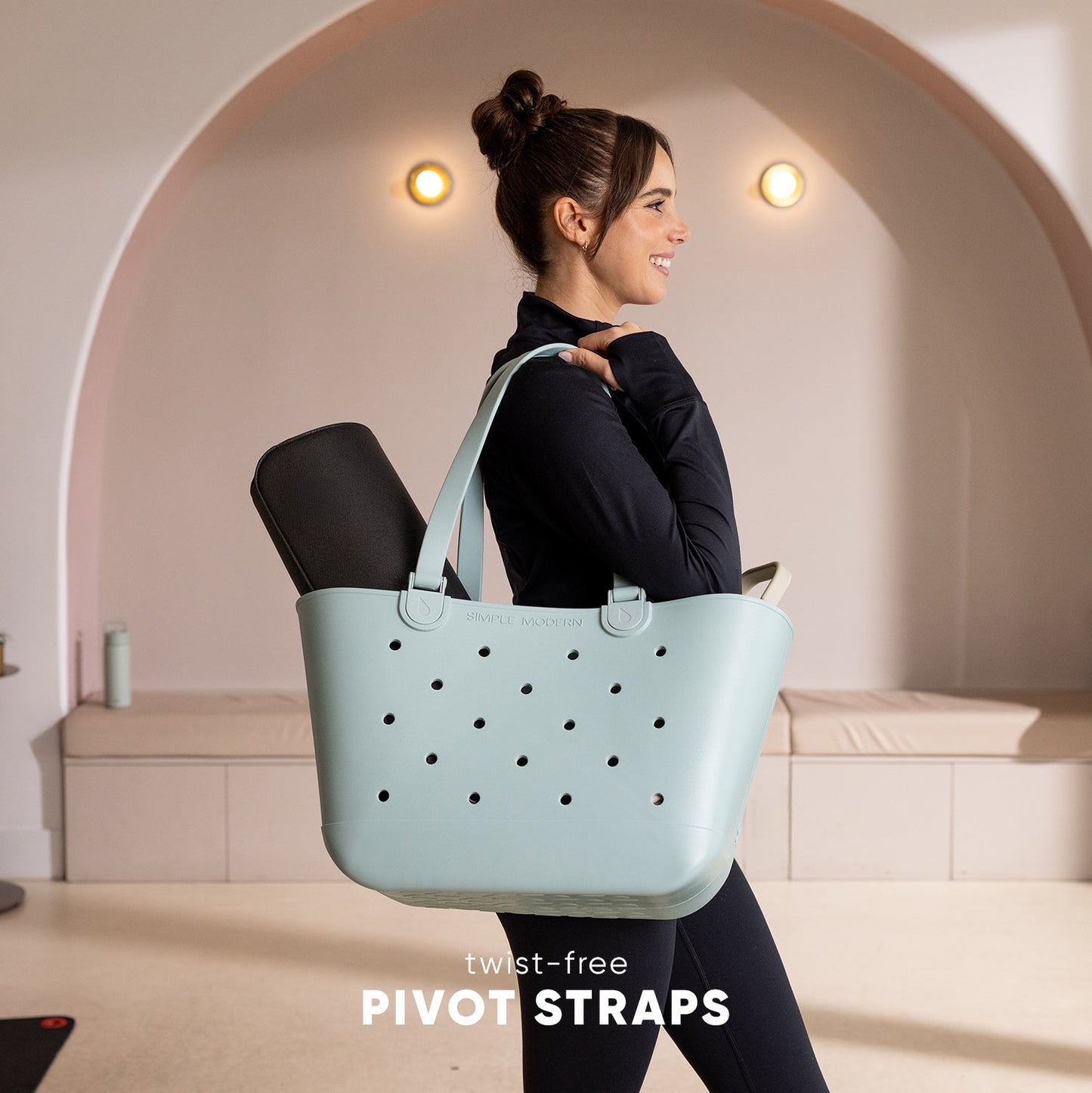 Woman wearing black top carrying light mint-green Simple Modern tote bag with perforated pattern and twist-free pivot straps in modern studio setting.