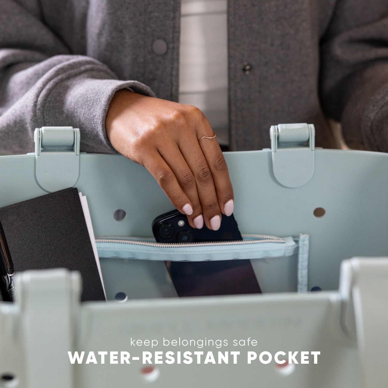 Woman placing black smartphone into water-resistant zippered pocket of light sage green Simple Modern tote bag with gray padded handles, demonstrating secure storage feature.