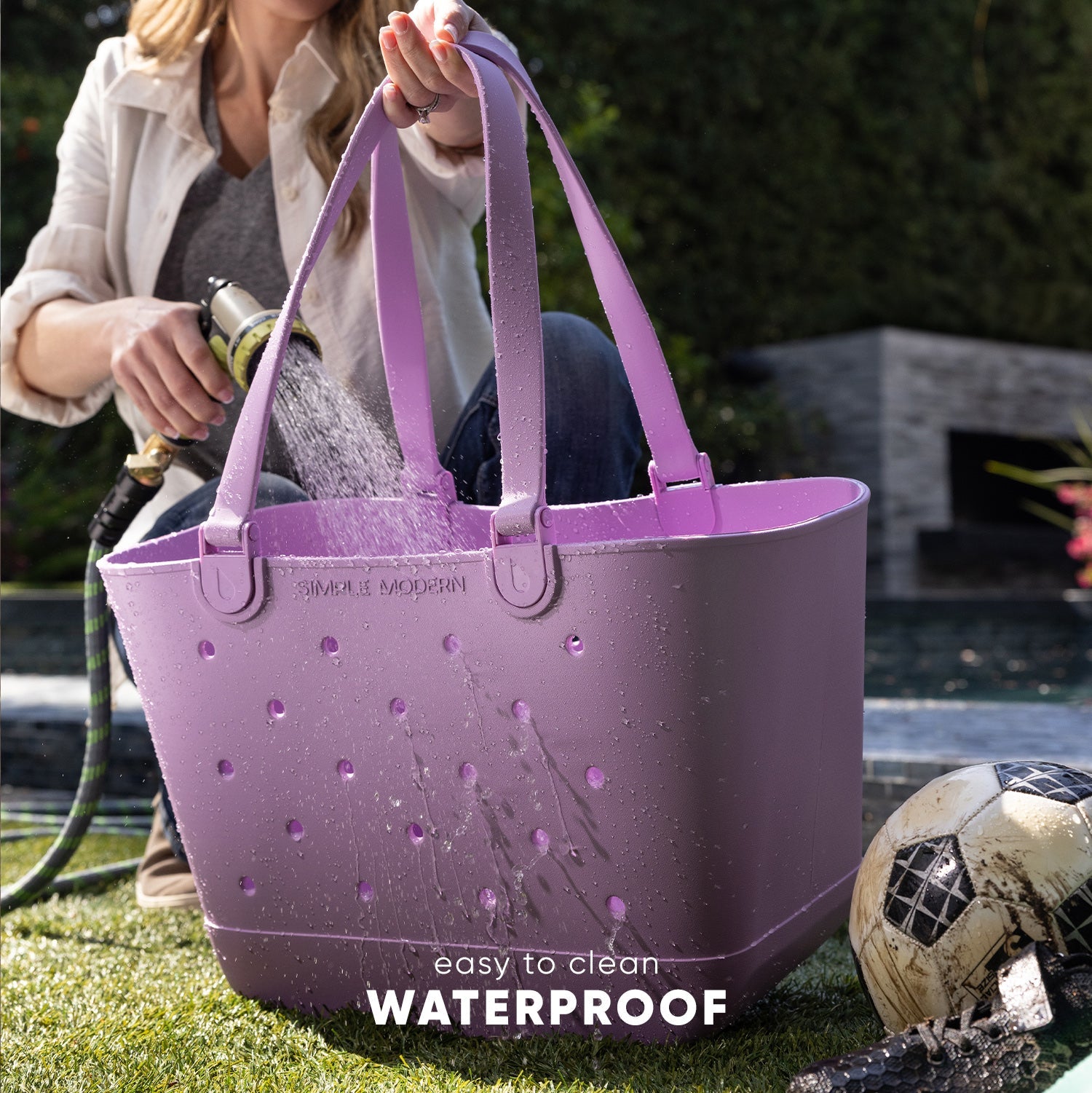 Lavender-purple waterproof getaway tote bag held by woman in garden, water beads visible on surface, "waterproof" and "easy to clean" text shown, garden setting with soccer ball and lawn visible.