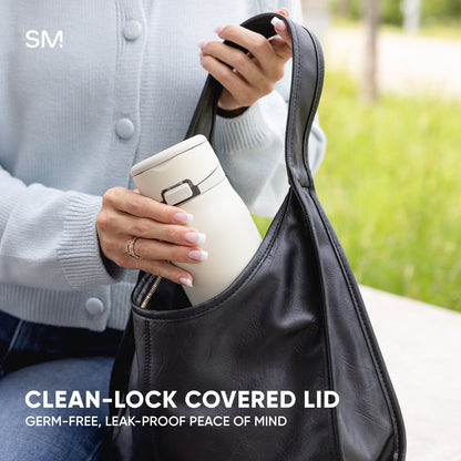 Woman holding cream-colored Simple Modern tumbler with metallic lock and covered lid, wearing gray cardigan and holding dark gray carrying strap, outdoor park setting with soft blurred green background.
