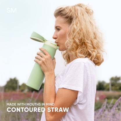 Woman in white t-shirt drinking from sage green SM water bottle with contoured straw in blooming flower field. Bottle features ergonomic design. Text reads "Made with Mouths in Mind, Contoured Straw."