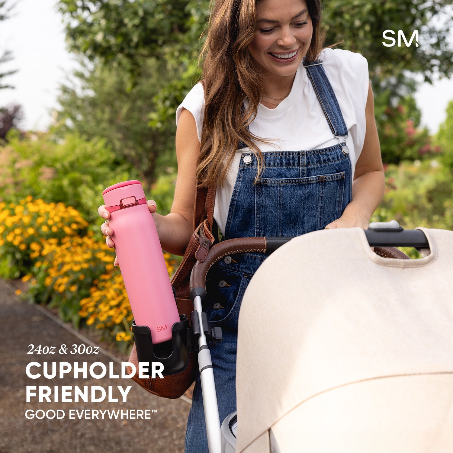 Woman in denim overalls holding a bright pink Simple Modern water bottle in a blooming garden with a cream stroller nearby. Text reads "24oz & 30oz Cupholder Friendly."