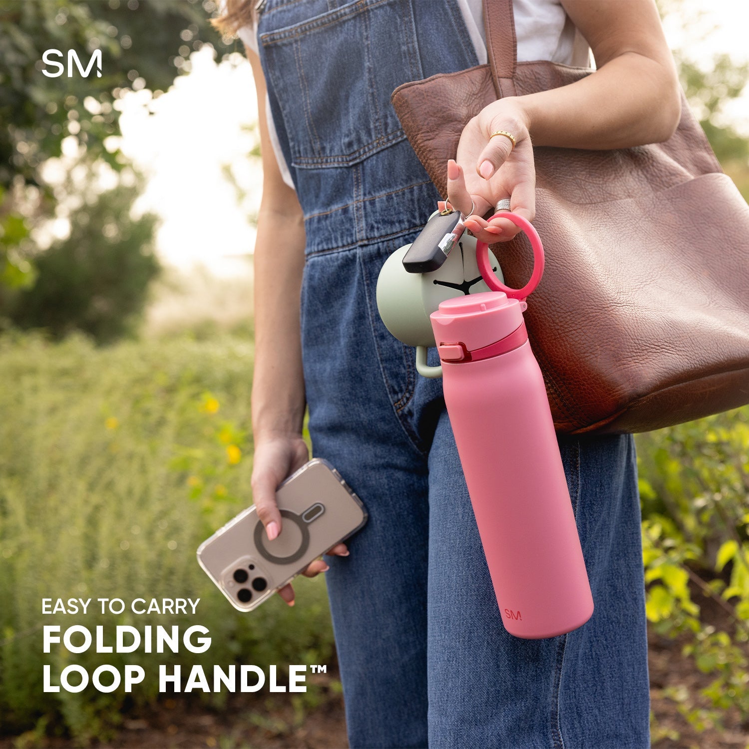 Woman holding a bright pink Simple Modern water bottle with black folding loop handle and red carabiner clip, paired with a brown leather shoulder bag, smartphone, and denim in an outdoor garden setting.