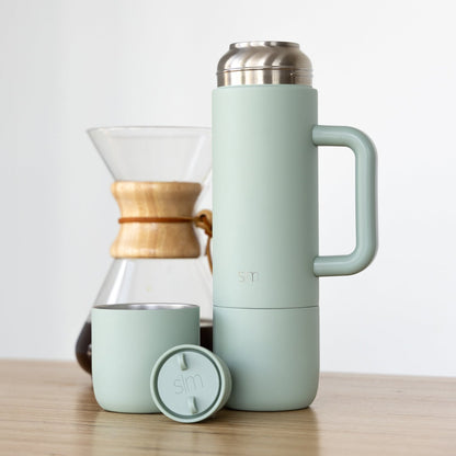 Sage green insulated tumbler and matching cup with warm wood and silver metallic accents on wooden surface