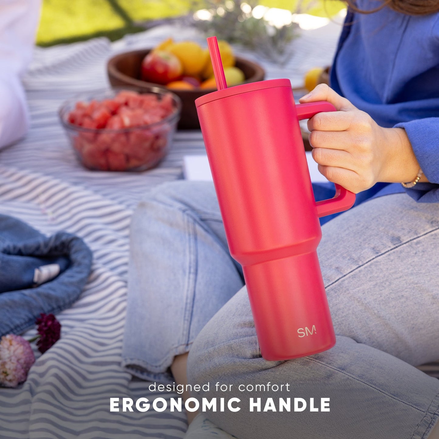 Coral pink insulated tumbler with ergonomic handle held by person in blue top during outdoor picnic, with fresh berries and fruits nearby on striped blanket. Text reads "designed for comfort" and "ERGONOMIC HANDLE."