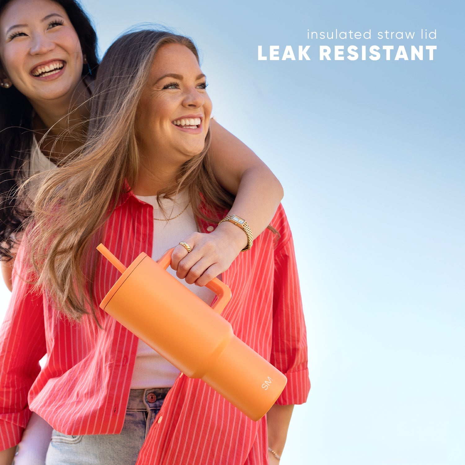 Two women smiling outdoors wearing red pinstriped blazers, holding a bright orange leak-resistant insulated tumbler with straw lid against a blue sky background.