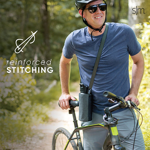 Man in blue-gray t-shirt and khaki pants standing with a sling-style bag across his body, holding a bicycle with black frame against green foliage background.
